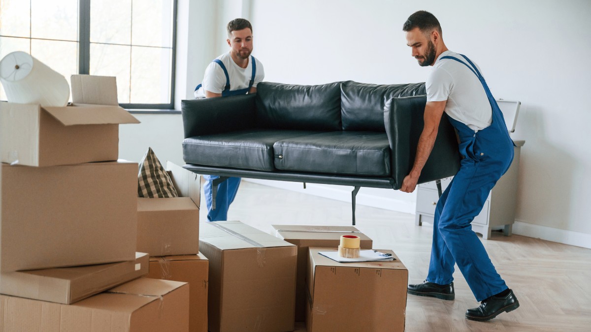 Two movers in blue overalls carry a black leather couch