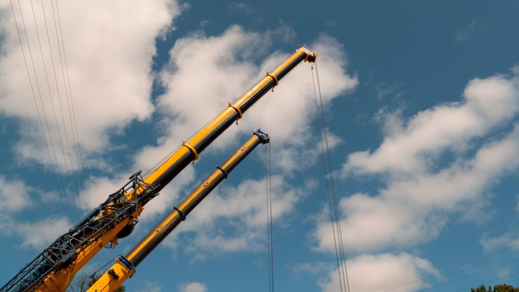 Two yellow construction cranes reaching upward against a clear blue sky with clouds.