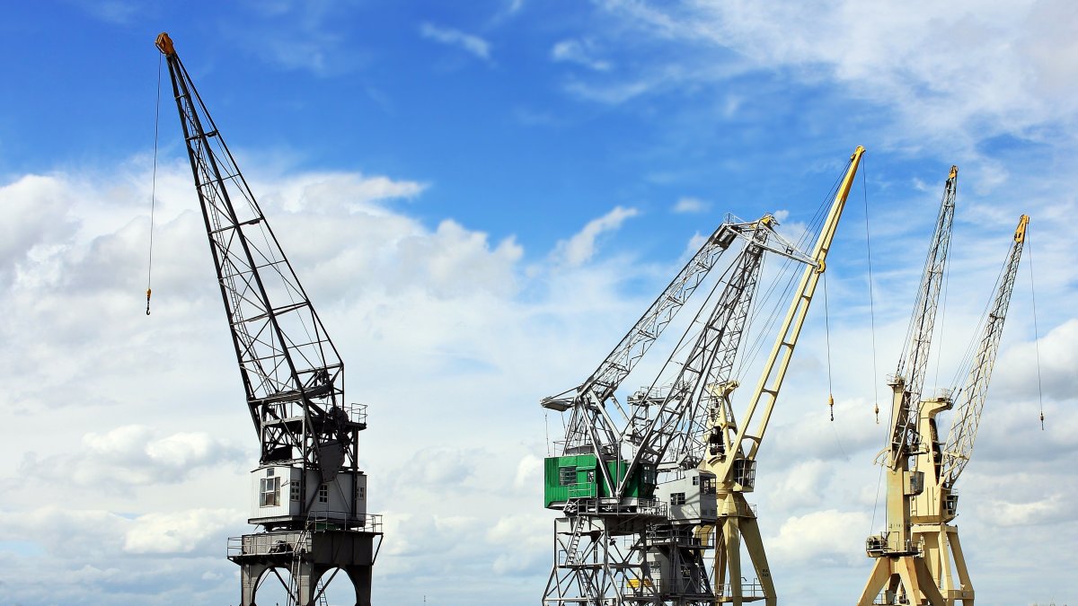 Industrial cranes reaching into a bright blue sky with scattered clouds, a symbol of construction and industry.